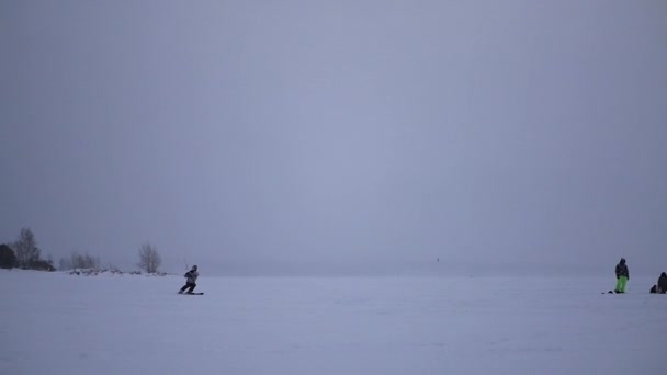 Sportowiec w kite zimowych, co skacze i ląduje na zamarzniętym jeziorze w śniegu w pobliżu brzegu, zwolnionym tempie — Wideo stockowe