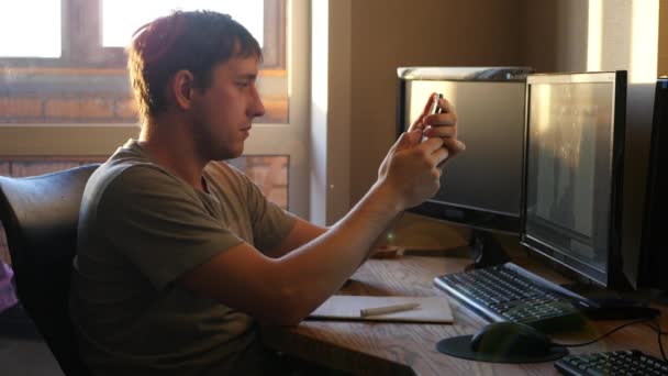 Ένας νεαρός άνδρας κάθεται στο γραφείο, στο ηλιοβασίλεμα, πάρτε το τηλέφωνο στο χέρι, γράφοντας κάτι ή να επιλέξετε το τηλέφωνο, 4k — Αρχείο Βίντεο