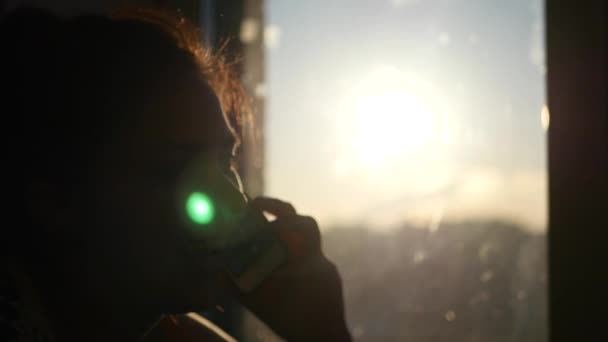 Schöne junge Mädchen spricht im Fenster auf dem Hintergrund Sonnenuntergang, 4k — Stockvideo