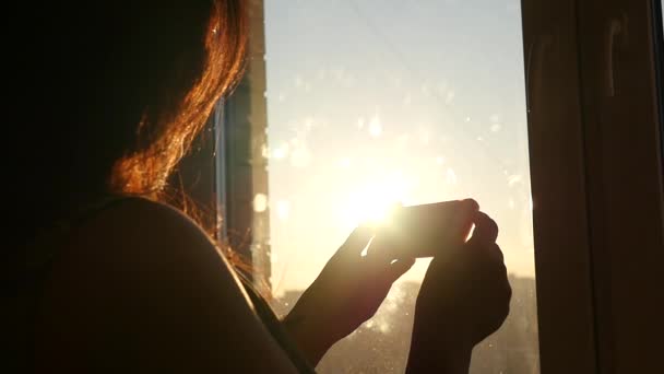 Joven hermosa chica conduce un dedo en el teléfono cerca de una ventana al atardecer, cámara lenta — Vídeos de Stock