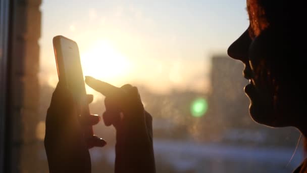 Mulher usa o telefone, dirige um dedo através da tela, olhando para o gadget, de pé na janela no fundo de um belo pôr do sol com brilho, 3840x2160, 4k — Vídeo de Stock