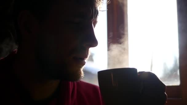 Tidigt på morgonen en man med ett skägg dricka varmt kaffe, är njuter av smaken mot fönstret, från muggen ett vackert par, Slowmotion — Stockvideo