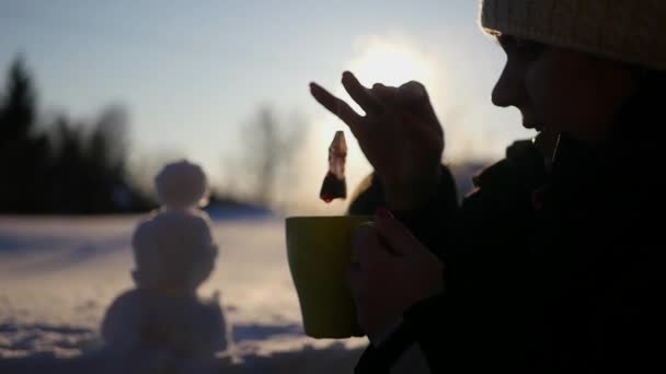 Ein Mädchen im Winter auf der Straße sitzt mit einer Tasse heißem Tee, brüht in einem Beutel, tropft in einen Wal, vor dem Hintergrund eines Sonnenuntergangs und eines Schneemanns. 1920x1080. Zeitlupe — Stockvideo