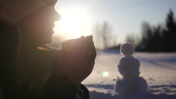 Een mooi meisje zit op straat met een kop dampende koffie, verwarmt handen, glimlacht, tegen de achtergrond van een sneeuwman en een gouden zonsondergang. 1920 x 1080. slow motion — Stockvideo