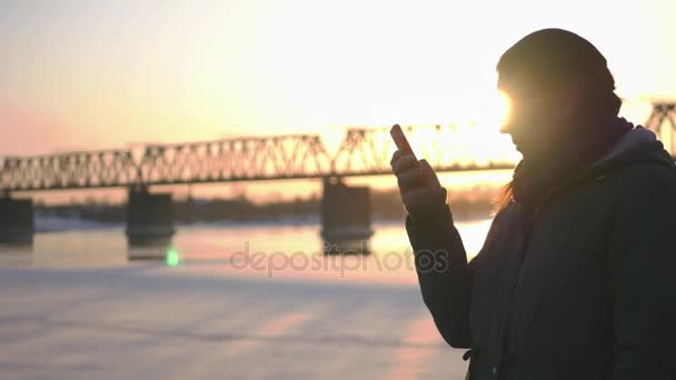 Een jong meisje loopt langs de kade op de achtergrond van een spoorbrug en een prachtige zonsondergang, bedrijf van een telefoon in haar handen, schrijft een bericht. 4k — Stockvideo