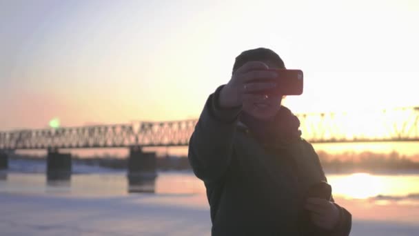 Une jolie fille se tient dans la rue et prend un selfie au coucher du soleil, et pont de fer. 4k — Video