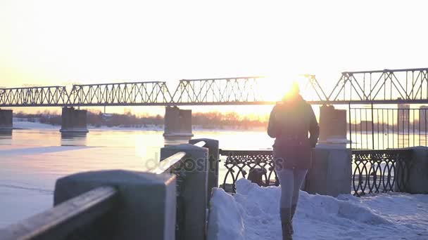 Yalnız bir kız nehir boyunca boyunca güzel sarı gün batımı ve demir köprü arka plan üzerinde yürür. HD — Stok video