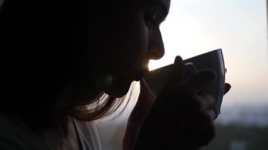 Genç çekici kız sıcak çay ya da güzel bir gün batımı ışınları bir lens etkisi ile bir pencerede oturup kahve içer. Yakın çekim. 1920 x 1080. ağır çekim. HD,