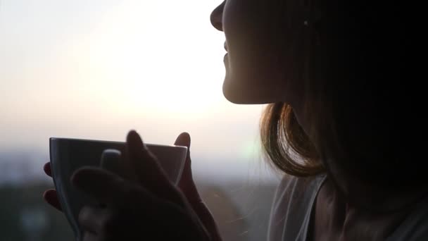 Au ralenti. Une belle jeune fille admire le coucher de soleil assis près de la fenêtre, boit une délicieuse boisson chaude dans une tasse blanche, des rayons de soleil sur son visage.. 1920x1080 — Video