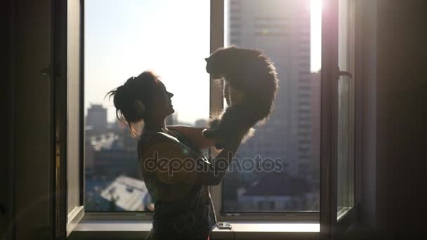 Hermosa chica en auriculares, hablando con un gato esponjoso en sus manos, contra el fondo de una ventana abierta con vistas a la ciudad de la noche, con una lente de efecto. En cámara lenta. 1920x1080 . — Vídeos de Stock