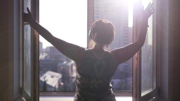 Una chica joven y hermosa se para cerca de una ventana y cierra las fajas, sobre un fondo de paisaje urbano con rayos de sol y el efecto de una lente. Slowmo 1920x1080 — Vídeo de stock