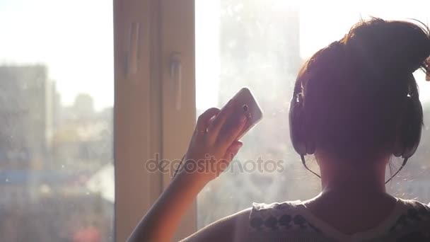 Veselá dívka v její ruce a sluchátka, střety hudby, tance, v paprscích slunce s efektem čočky stojí u okna s telefonem. Slowmo 1920 × 1080 — Stock video