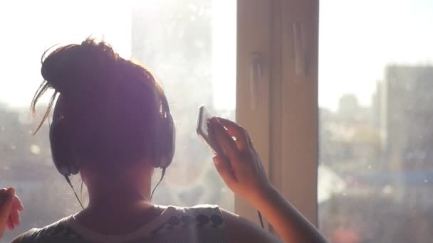Дівчина у великих навушниках танцює біля вікна на міському фоні, сонячні промені з телефоном в руках. Повільна 1920x1080 — стокове відео
