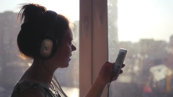 Jovencita adolescente en auriculares canta una canción, mira en el teléfono en un fondo del paisaje de la ciudad fuera de la ventana. En cámara lenta. 1920x1080 . — Vídeos de Stock