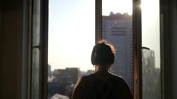 Una joven que está de pie en la ventana abierta disfruta del sol de primavera en la ciudad, con el efecto de una lente. En cámara lenta. 1920x1080 . — Vídeos de Stock