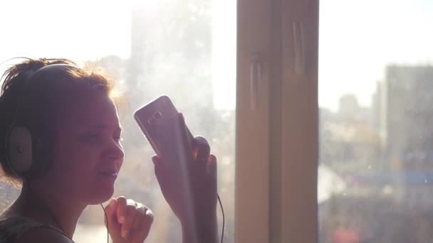 Веселая девушка в нуашниках танцует, слушает музыку по телефону. Смотрит в камеру, улыбаясь, на фоне городского пейзажа за окном. Слоумо 1920x1080 — стоковое видео