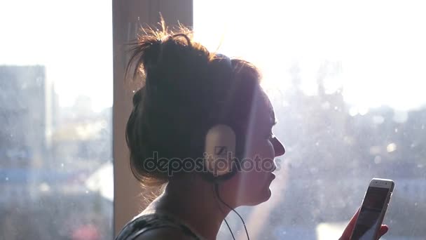 Молодая веселая девушка в наушниках и с телефоном в руках танцует и поет, стоя у окна под утренним солнцем. Медленное движение. 1920x1080 . — стоковое видео