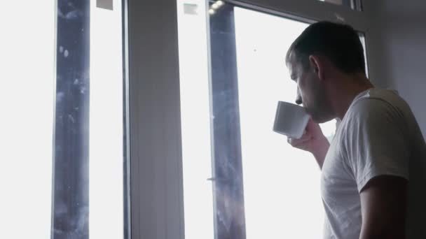 Young attractive man in white t-shirt drinking coffee standing by the window, looking to the distance. 4k, 3840x2160. HD — Stock Video