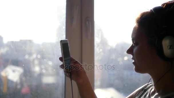 Egy fiatal lány áll az első fejhallgató és a telefon a kezében, egy ablak néz a szerkentyű. Slowmotion. 1920 x 1080 — Stock videók