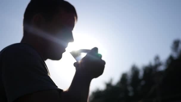 Un jeune homme en bonne santé boit de l'eau propre à partir d'un gobelet de verre, debout contre le soleil de profil, dans les rayons et l'effet de la lentille. En Haute Qualité, 1920x1080 . — Video