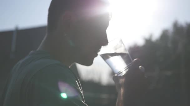 Um jovem bebe água limpa de um copo, gotas de água fluem ao longo do fundo e brilham nos raios do sol. Motiona lenta. Full HD 1920x1080p — Vídeo de Stock