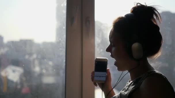 Fille gaie dans de gros écouteurs dansant près d'une fenêtre sur un fond de ville, les rayons du soleil avec un téléphone dans les mains. Slowmo 1920x1080 — Video