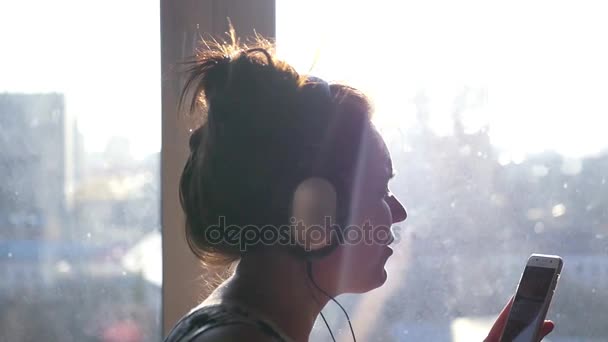 En glad flicka i hörlurar och med en telefon i hennes händer dansar och sjunger stående nära fönstret i morgonsolen. Slow motion. 1920 x 1080. — Stockvideo
