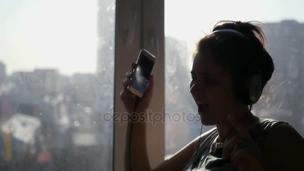 Una joven alegre escucha música en los auriculares desde el teléfono, canta y baila en voz alta junto a la ventana al sol. cámara lenta. 1920x1080 — Vídeo de stock