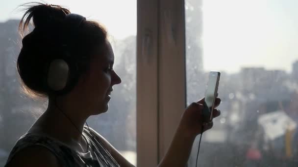 Uma jovem menina bonita fica perto de uma janela em seus fones de ouvido e vira através da lista de reprodução de telefone. câmara lenta. 1920x1080 — Vídeo de Stock