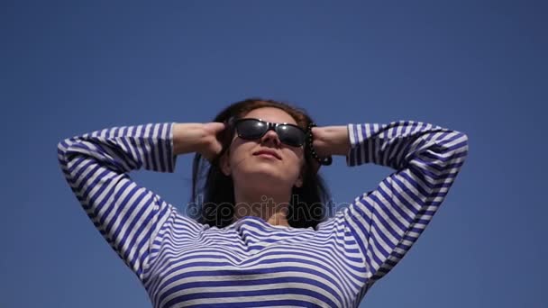 Giovane bella ragazza gode il sole e il calore in una camicetta a righe, svolazzando per i capelli contro un cielo blu chiaro. Video al rallentatore 1080p full HD . — Video Stock