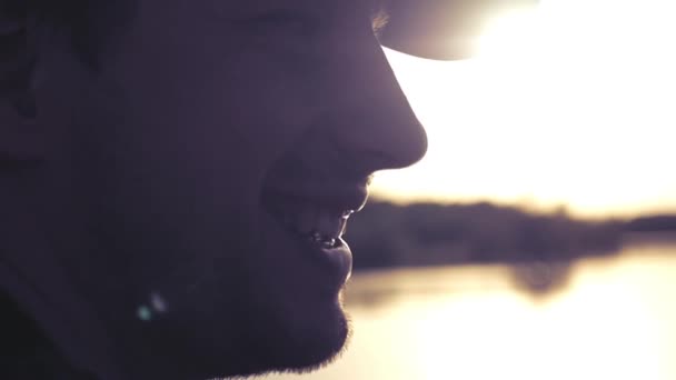 Vuxen man i baseball cap sitter i profil på sjön, tittar på solnedgången, leende. slowmotion. 1920 x 1080 — Stockvideo