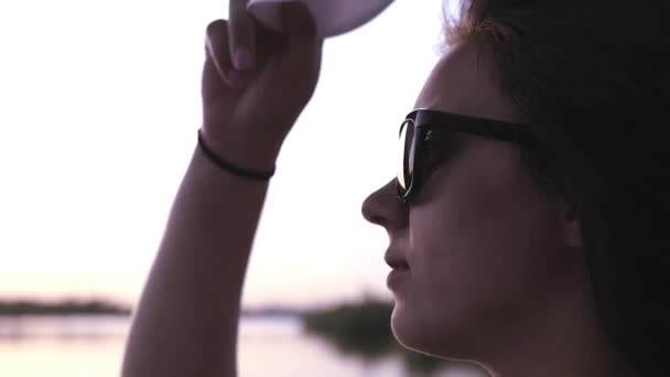 A young beautiful girl stands on the seashore admires the sunset in sunglasses, puts on a cap. slow motion. 1920x1080 — Stock Video