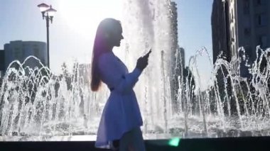 Güzel ince kız standları bir telefonla onun elinde çeşmenin bir ileti yazıyor telefon yaz güneş ışınlarının yansıtır. Ağır çekim. HD, 1920 x 1080.