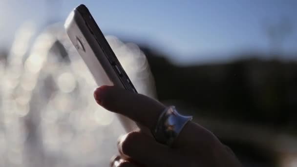 Стильный смартфон в руках девушки, блестит на солнце, с фонтаном на заднем плане в размытом медленном движении. HD, 1920x1080 . — стоковое видео