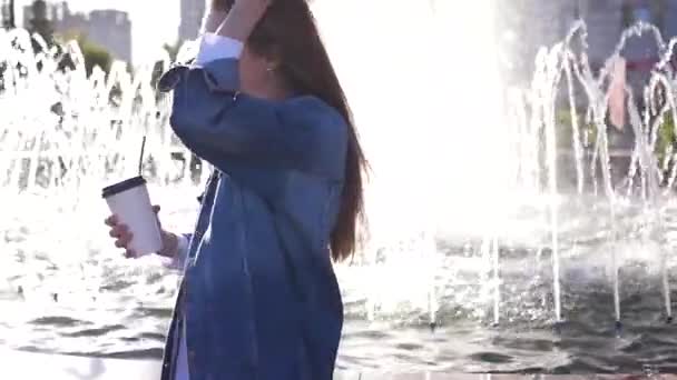Una hermosa chica elegante bebe café de una taza de papel a través de un doblaje, de pie junto a la fuente con la luz del sol y la reflexión. Lento movimiento. HD, 1920x1080 . — Vídeos de Stock