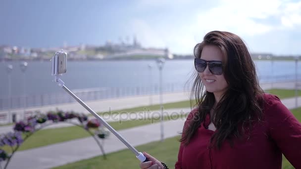 Giovane bella ragazza sta facendo una foto su un bastone selfie su uno sfondo di acqua, città e cielo. HD, 1920x1080. Rallentatore — Video Stock