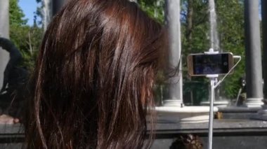 Genç kız bir resmi telefon ve selfie sopa için parkta, görünümü bir çeşme bir arka plan üzerinde arkadan yapar. ağır çekim. 1920 x 1080. hd tam