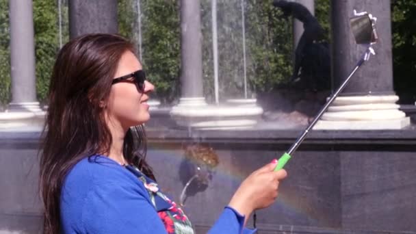 Девушка делает фото по телефону с помощью селфи-палки на фоне фонтана и красивой радуги. Замедление. 1920x1080. полный комплект — стоковое видео