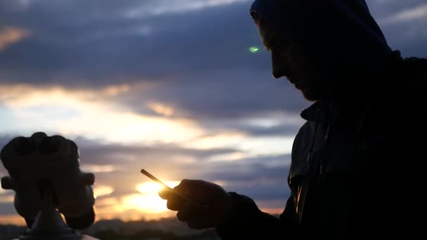 Mann schaut in das Telefon auf Sonnenuntergang und Teleskophintergrund. Zeitlupe, 1920x1080, full hd — Stockvideo