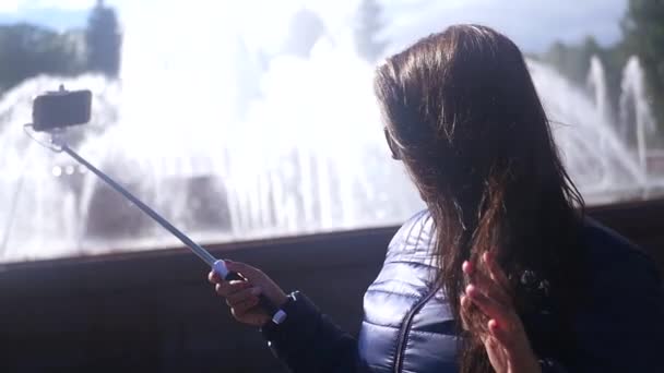 La chica hace una foto en el palo selfie, cerca de la fuente, el pelo se desarrolla a favor del viento. cámara lenta. 1920x1080. hd completa — Vídeo de stock