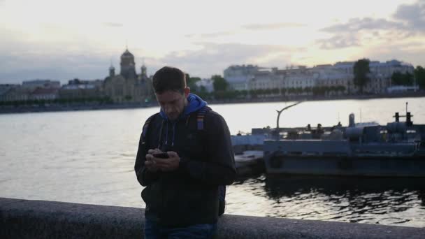 A young man stands on the embankment of a river with a telephone, writes a message, against the backdrop of a sunset, the city and the church. slow motion. 1920x1080. full hd — Stock Video