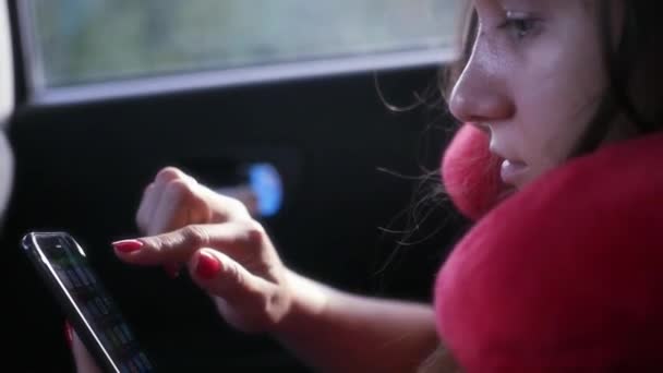 Chica pasajero viaja en el coche en el asiento trasero, con una cómoda almohada debajo del cuello, sostiene el teléfono en las manos, presiona la pantalla. HD, 1920x1080. cámara lenta — Vídeos de Stock