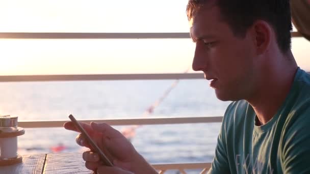 A young attractive man is sitting with a phone in his hands at sunset near the sea, is driving his finger across the screen. HD, 1920x1080. slow motion