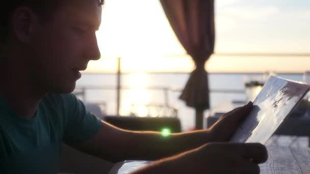 Un jeune homme assis dans un café au bord de la mer avec Zakia, pouce au menu, veut faire un choix. HD, 1920 x 1080. au ralenti — Video