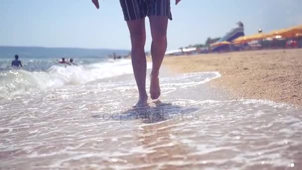 Un hombre está caminando a lo largo de una playa de arena, las olas se rompen alrededor de sus pies. HD, 1920x1080. cámara lenta — Vídeo de stock