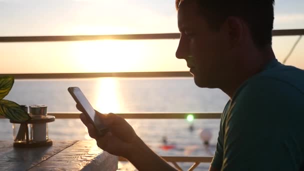 Um homem senta-se em um café no fundo de um belo pôr do sol, com um telefone em suas mãos, escreve uma mensagem. HD, 1920x1080. câmara lenta — Vídeo de Stock