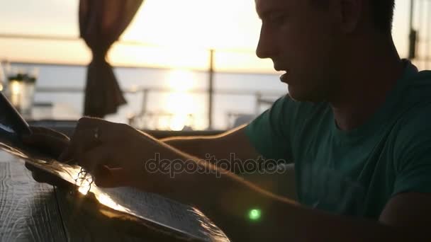 Un joven se sienta en un café y selecciona la comida del menú, sobre el fondo de una hermosa puesta de sol en el horizonte. HD, 1920x1080. cámara lenta — Vídeo de stock