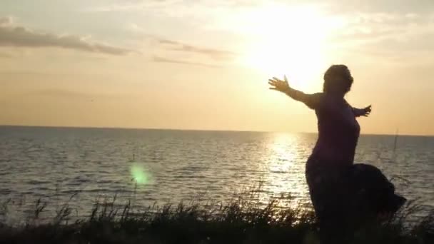 Ein junges Mädchen dreht sich im Gras vor dem Hintergrund des Sonnenuntergangs und des Meeres und genießt die Sonne. 4k, 3840x2160. hd — Stockvideo