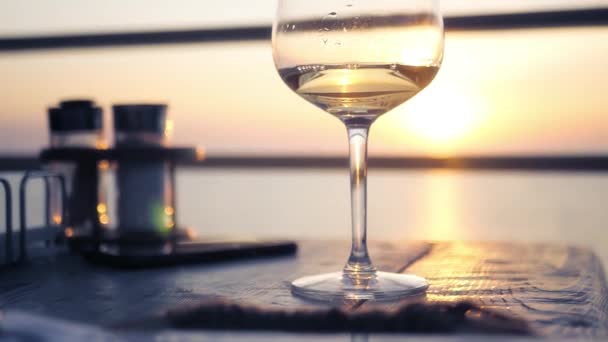 На столі на фоні заходу сонця і моря на горизонті стоїть келих з білим вином. HD, 1920x1080. повільний рух . — стокове відео