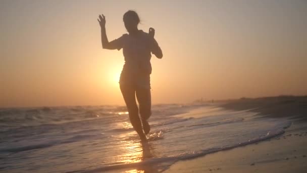 Una chica delgada y alegre corre a lo largo de las olas en salpicaduras de agua al atardecer. HD, 1920x1080. cámara lenta . — Vídeo de stock